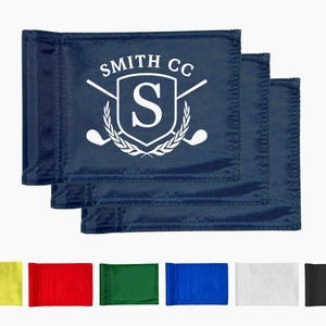 May include: Navy blue golf flags with the text "SMITH CC" and a white logo featuring a shield, the letter "S", crossed golf clubs, and a laurel wreath. Additional flags in yellow, red, green, blue, white, and black are shown.