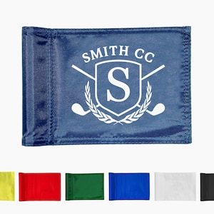 May include: A dark blue golf flag featuring a white printed design of a shield with the letter S, crossed golf clubs, and a laurel wreath, with the text SMITH CC arched above. Below are six additional golf flags in yellow, red, green, blue, white, and black.
