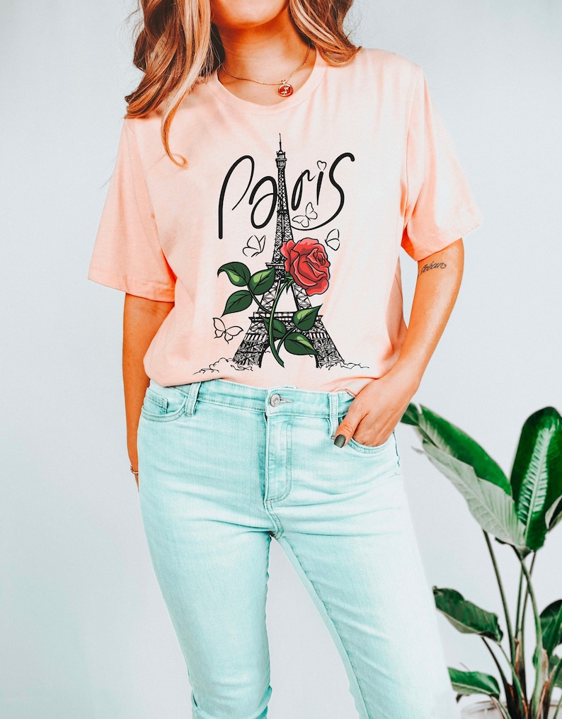 May include: A pink t-shirt with a black and white graphic of the Eiffel Tower with a red rose and butterflies. The text 'Paris' is written in black cursive above the tower. The t-shirt is being worn with light blue jeans.