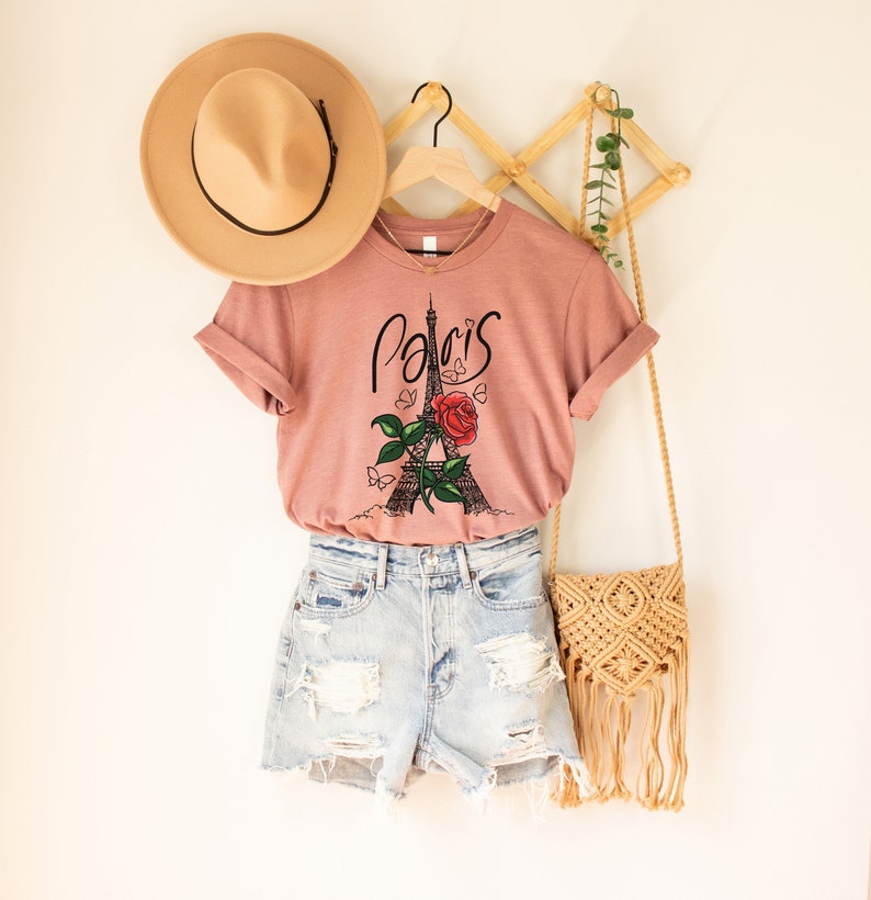 May include: A dusty rose t-shirt with a graphic of the Eiffel Tower and a red rose. The text "Paris" is written in cursive above the tower. The t-shirt is hanging on a wooden hanger.