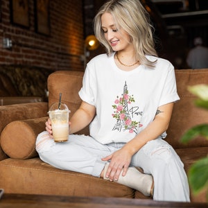 May include: A woman wearing a white t-shirt with a black and pink Eiffel Tower graphic and the word "Paris" on it. She is sitting on a brown couch and holding a iced coffee.