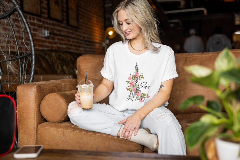 May include: A woman wearing a white t-shirt with a black and pink Eiffel Tower graphic and the word "Paris" on it. She is sitting on a brown couch and holding a iced coffee.