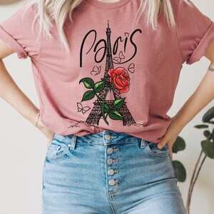 May include: A pink t-shirt with a graphic of the Eiffel Tower, a red rose, and butterflies. The text "Paris" is written in a cursive font above the tower.