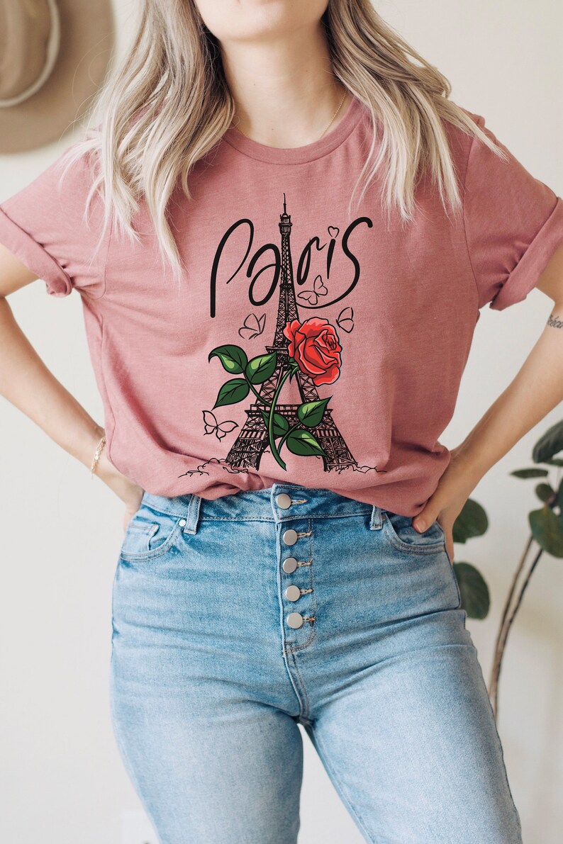 May include: A pink t-shirt with a graphic of the Eiffel Tower, a red rose, and butterflies. The text "Paris" is written in a cursive font above the tower.