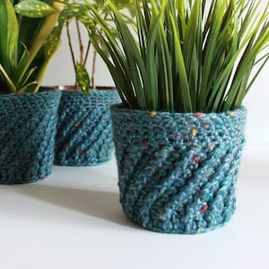 Puede incluir: Tres macetas de plantas de color azul turquesa tejidas a crochet con diferentes tipos de plantas verdes. Las macetas tienen un diseño texturizado y tejido.
