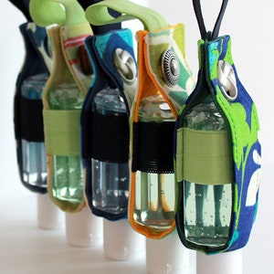 May include: Five colourful fabric hand sanitiser holders with clear bottles of hand sanitiser inside. The holders have a loop at the top for carrying.