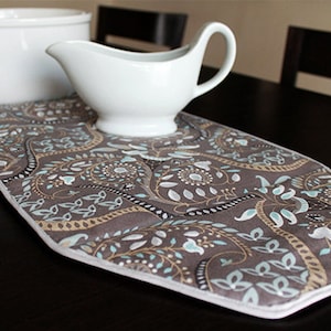 May include: A brown and white paisley patterned table runner with a white ceramic gravy boat on top.