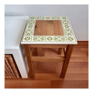 Op de afbeelding: Een kleine houten bijzettafel met een wit blad en een groen bloemenstencilontwerp. De tafel heeft één plank en is gemaakt van natuurlijk hout.