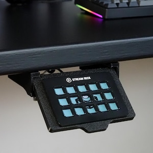 May include: A black Stream Deck controller mounted underneath a desk. The device features a grid of fifteen square buttons with light blue illuminated icons. The controller is held in place by a black plastic mounting bracket attached to the desk.
