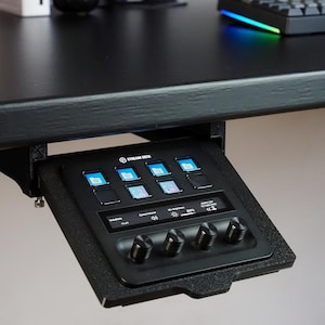 May include: A black Stream Deck controller mounted under a desk, featuring eight square LCD keys with blue folder icons, a horizontal touch display showing system volume and brightness settings, and four circular black control knobs at the bottom.