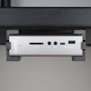 Könnte beinhalten: Eine silberne CalDigit-Dockingstation in einer schwarzen Kunststoffhalterung. Die Vorderseite verfügt über einen SD-Kartenslot, zwei 3,5-mm-Audioanschlüsse, einen USB-C-Anschluss und einen USB-A-Anschluss mit der Beschriftung USB 5Gbps sowie eine Statusleuchte.