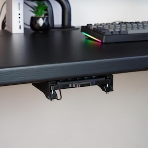 May include: A black under-desk mount bracket attached to the underside of a desk. The mount features a rigid frame with two vertical support arms and metal hardware, designed to hold a device securely beneath the flat surface of the desk.