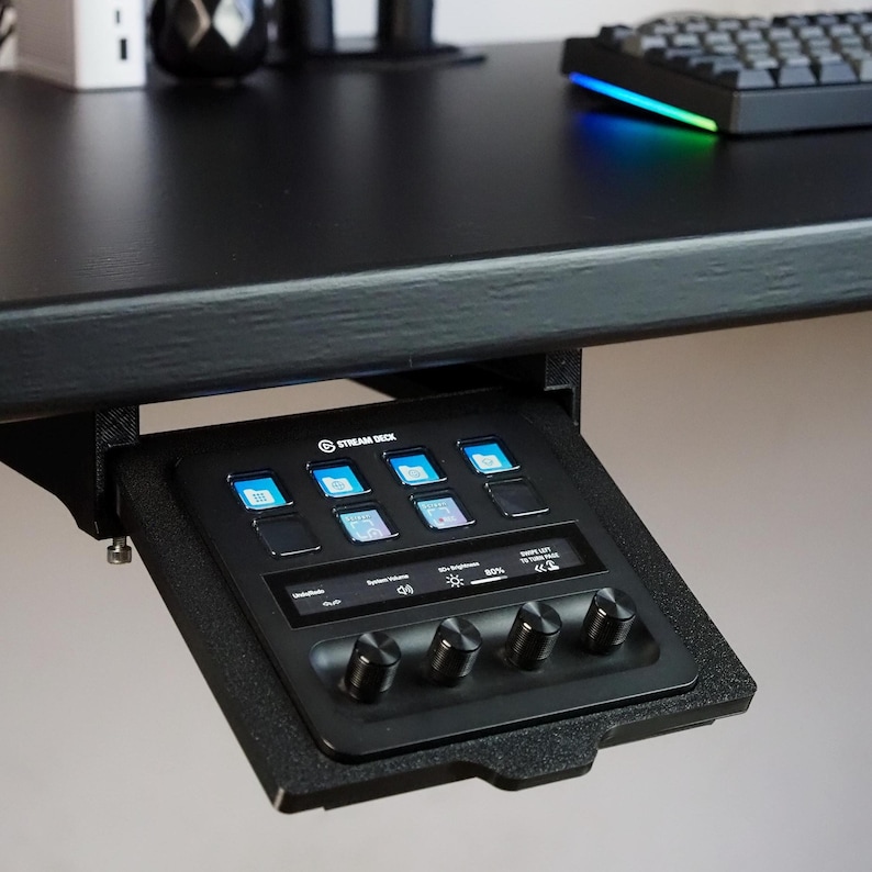 May include: A black Stream Deck controller mounted under a desk, featuring eight square LCD keys with blue folder icons, a horizontal touch display showing system volume and brightness settings, and four circular black control knobs at the bottom.