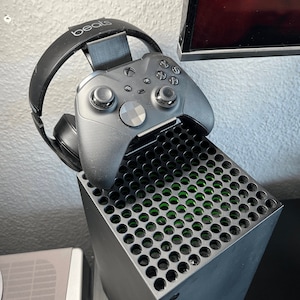 Könnte beinhalten: Eine schwarze Xbox Series X Konsole mit einem schwarzen und grünen Lüftungsgitter auf der Oberseite. Ein schwarzer Xbox Controller ist an einem schwarzen Beats by Dre Kopfhörerständer befestigt.