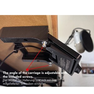 May include: A black 3D-printed mounting bracket attached to the underside of a wooden desk, featuring an adjustable carriage mechanism secured with metal screws. The text reads: The angle of the carriage is adjustable with the included screws.