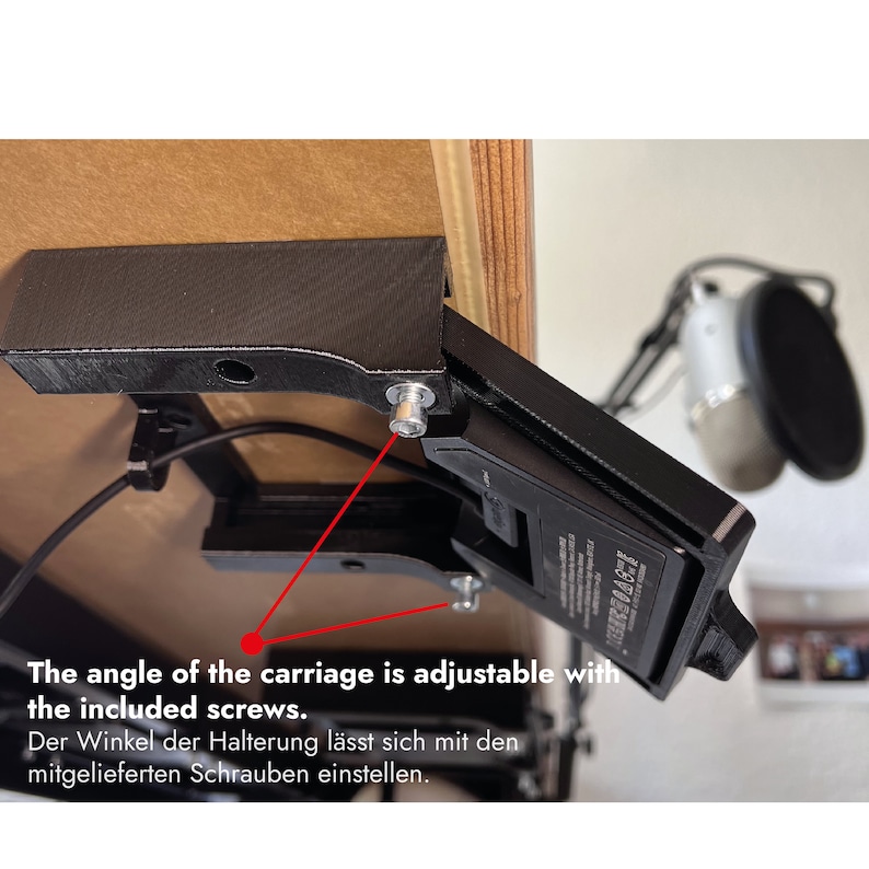 May include: A black 3D-printed mounting bracket attached to the underside of a wooden desk, featuring an adjustable carriage mechanism secured with metal screws. The text reads: The angle of the carriage is adjustable with the included screws.