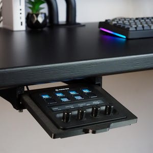 May include: A black Stream Deck controller mounted under a desk, featuring eight square LCD keys with blue icons and four circular rotary knobs. The device is housed in a black 3D-printed bracket attached to the underside of a black desk surface.