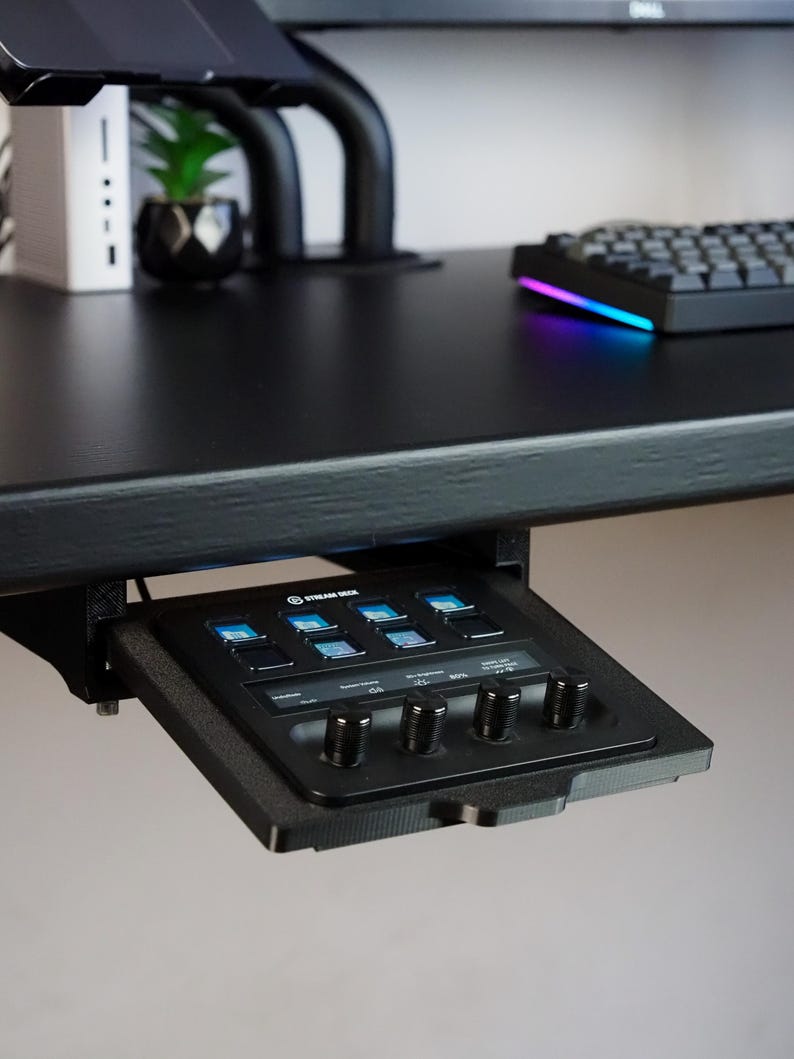 May include: A black Stream Deck controller mounted under a desk, featuring eight square LCD keys with blue icons and four circular rotary knobs. The device is housed in a black 3D-printed bracket attached to the underside of a black desk surface.