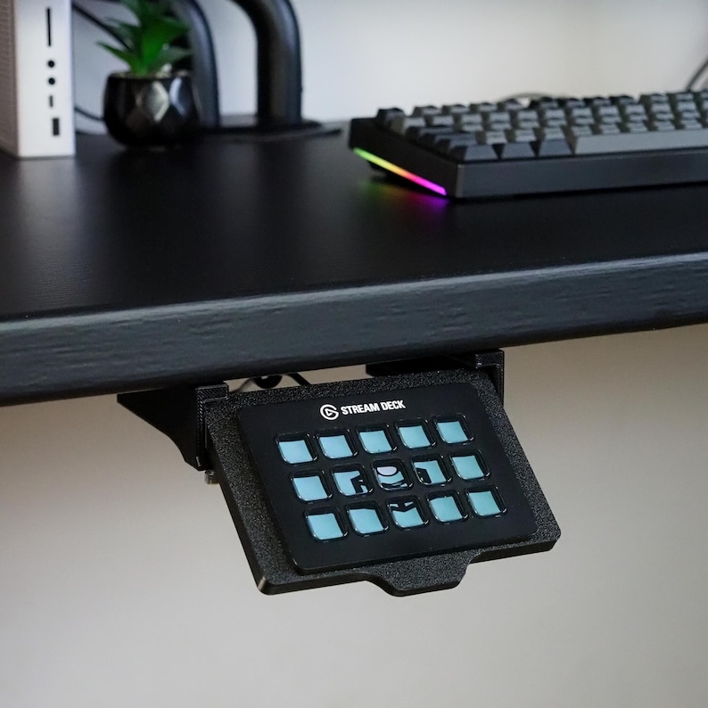 May include: A black Stream Deck controller mounted underneath a desk. The device features a grid of fifteen square buttons with light blue illuminated icons. The controller is held in place by a black plastic mounting bracket attached to the desk.