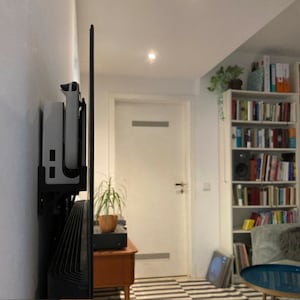 May include: A white video game console is mounted on a wall next to a black television screen. The console is attached to a black wall mount. The console is in a living room with a bookshelf and a door in the background.