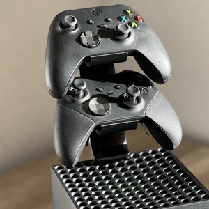 Könnte beinhalten: Zwei schwarze Xbox-Controller sind auf einem Ladeständer gestapelt, der auf einer schwarzen Xbox Series X-Konsole steht.