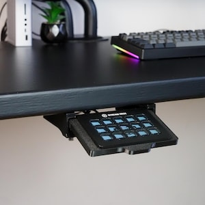May include: A black Stream Deck controller mounted underneath a black desk surface using a custom 3D-printed bracket. The device features fifteen square buttons with blue illuminated icons and the Stream Deck logo printed in white at the top center.