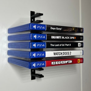 Puede incluir: Una estantería de pared de metal negro que contiene cinco cajas de juegos de PS4. Los títulos de los juegos visibles son "Days Gone", "Call of Duty: Black Ops", "The Last of Us Part II", "Watch Dogs 2" y "Red Dead Redemption II". Las cajas son azules y negras.
