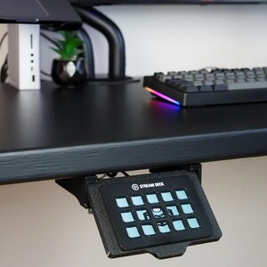 May include: A black Stream Deck controller mounted underneath a desk edge. The device features a grid of twelve square buttons with light blue illuminated icons and the text STREAM DECK printed in white at the top center of the black frame.