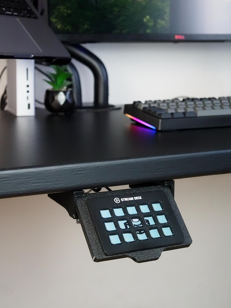 May include: A black Stream Deck controller mounted underneath a desk edge. The device features a grid of twelve square buttons with light blue illuminated icons and the text STREAM DECK printed in white at the top center of the black frame.