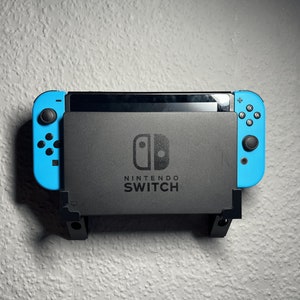May include: A blue and black Nintendo Switch console mounted on a wall with a black wall mount. The console has the Nintendo Switch logo and the words "Nintendo Switch" printed on it.