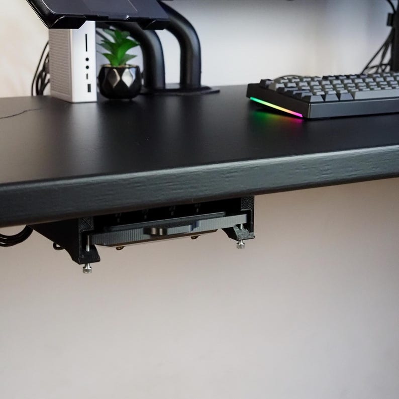 May include: A black under-desk mount attached to the underside of a black desk surface. The mount features a rectangular frame with visible metal screws and holds a flat, thin electronic device securely in place beneath the workspace.