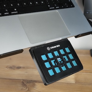Könnte beinhalten: Ein schwarzes Stream Deck-Bedienfeld mit sechzehn beleuchteten blauen Tasten befindet sich unter einem silbernen Laptop. Das Stream Deck-Logo ist sichtbar. Das Gerät befindet sich auf einer Holzoberfläche.