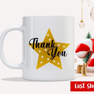 May include: White ceramic mug featuring a gold star design with the words "Thank You" in black script. The star is filled with small white stars. The mug has a curved handle and is set against a festive background.