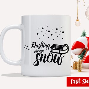 以下が含まれることがあります： 白いセラミック製のマグカップに、黒いデザインで「Dashing through snow」と書いてあり、そりのシルエットが描かれています。