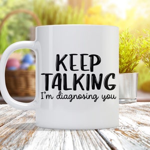May include: White ceramic coffee mug with black text that reads "Keep Talking I'm diagnosing you".