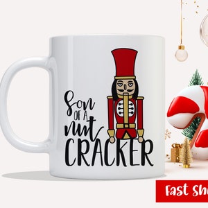 May include: White ceramic mug with a black and red illustration of a nutcracker and the text "Son of a nut cracker".
