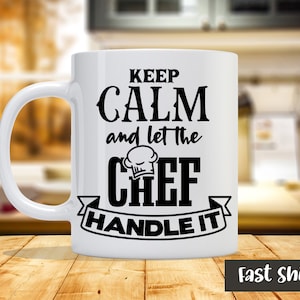 Könnte beinhalten: Weiße Keramiktasse mit schwarzem Text, der "Keep Calm and Let the Chef Handle It" lautet. Der Text ist in einer stilisierten Schriftart mit einer Kochmütze über dem Wort "Chef".
