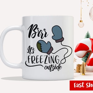 May include: White ceramic mug with a black and blue illustration of mittens and the text "Brrr It's Freezing Outside".