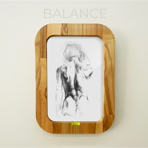 May include: A framed drawing of a nude figure in a standing pose. The drawing is in black and white and is framed in a light wood frame. The frame has the word "BALANCE" printed on the top.