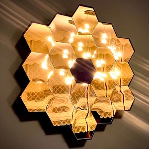 Op de afbeelding: Een decoratieve wandspiegel bestaande uit meerdere zeshoekige spiegeltegels. De spiegel reflecteert een warm, goudkleurig licht en de afbeelding van hangende hanglampen. Het frame is donker en de spiegel is op een grijze muur gemonteerd.