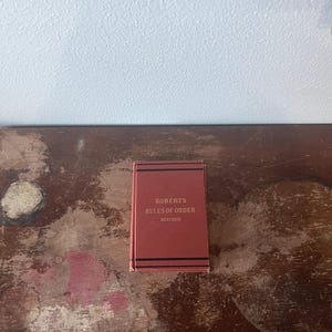 May include: A small, rectangular book titled "Robert's Rules of Order Revised" in gold lettering on a red cover. The book has black horizontal stripes and is set against a distressed wooden surface.