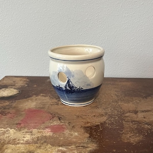May include: A ceramic onion keeper with a cream-colored base and a blue nautical scene. The keeper has two circular holes and a blue rim. The design features a sailboat on the sea.