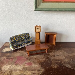 May include: Miniature dollhouse furniture set including a sofa with a blue and yellow floral pattern, a wooden coffee table, a grandfather clock, and a small side table. The furniture is arranged on a distressed wooden surface.