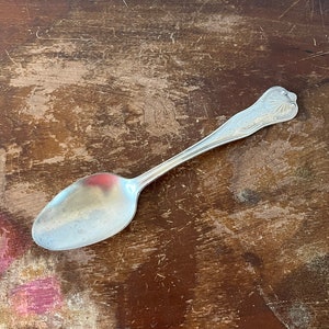 May include: A silver spoon with an ornate handle on a brown wooden surface.