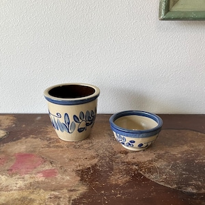 May include: Two ceramic pieces: a small bowl and a cylindrical pot. Both are beige with blue leaf patterns and a blue rim. The pot has a dark brown interior. The items are on a distressed wooden surface.