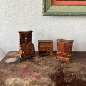 May include: Three miniature wooden furniture pieces, including a secretary desk, a hutch with glass doors, and a cabinet with two doors. The pieces are all brown and have a distressed finish.