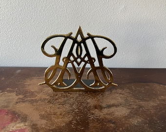 May include: A gold metal napkin holder with a decorative design featuring the letter "A".