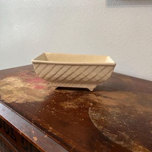 May include: A small, cream-coloured ceramic planter with a woven design and four feet. The planter is sitting on a wooden surface.