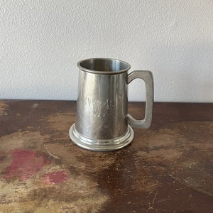 May include: A silver pewter tankard with a rounded base and a handle. The tankard has an inscription on the front.
