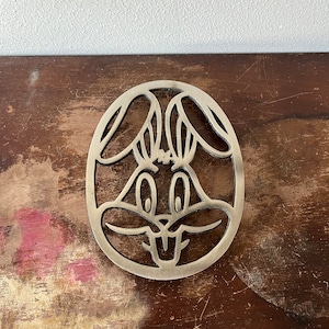 May include: A silver metal trivet shaped like a cartoon rabbit's face. The trivet has a cutout design and is perfect for protecting surfaces from hot dishes.